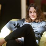 A young woman with short brown hair and a black long-sleeved boiler suit sits in a green armchair with her legs crossed. Dappled sunlight. She looks at something off camera and smirks.