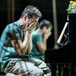 Andrew Scott is wearing a green shirt, sitting on a piano stool, his face covered by his hands. A black piano on which a bunch of yellow and white flowers is placed, is in front of him.