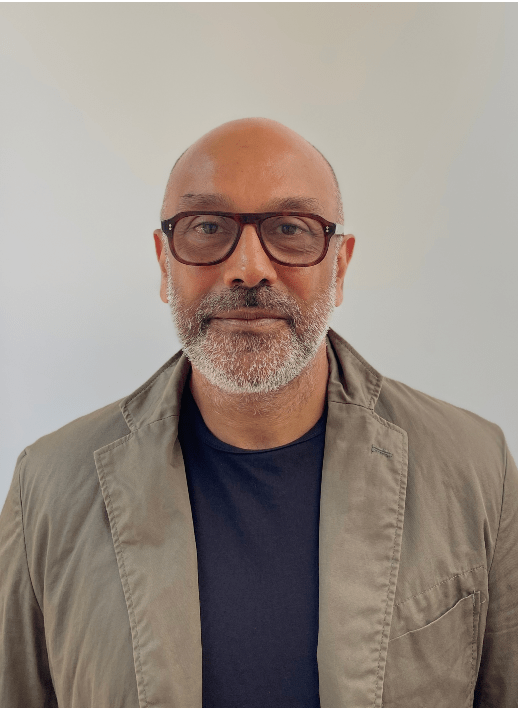 A photograph of a person wearing glasses, with a grey-ish beard and wearing a beige jacket and navy blue t-shirt