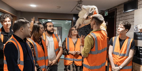 A group of people in high vis orange jackets stand and listen to a man that holds a puppet of a polar bear head on his hand.